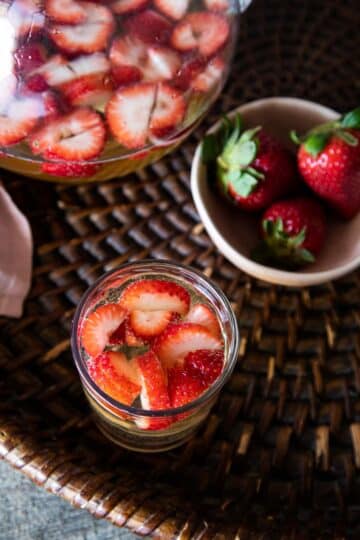 Glass filled with sangria and fresh strawberry slices, beside a bowl with more strawberries.
