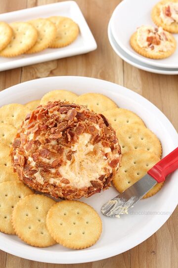 Cheese ball covered in bacon, on a serving plate with crackers and a knife.