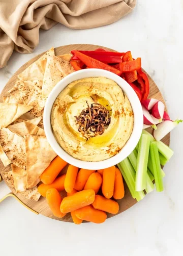 A bowl of hummus on a serving tray surrounded by fresh veggies and pita chips.