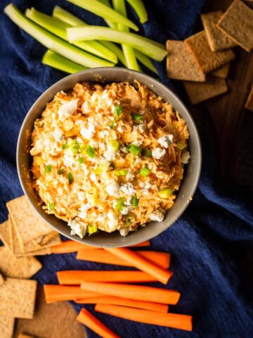 A serving bowl filled with cold buffalo chicken dip served with veggies.