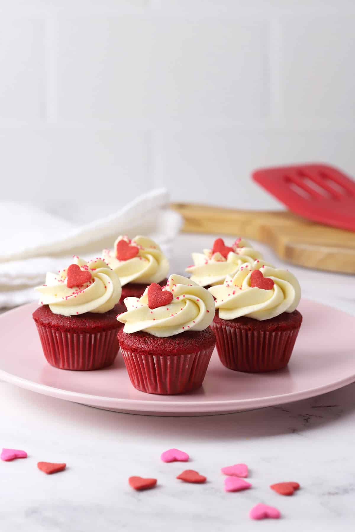 A pink plate filled with mini red velvet cupcakes topped with heart shaped sprinkles.