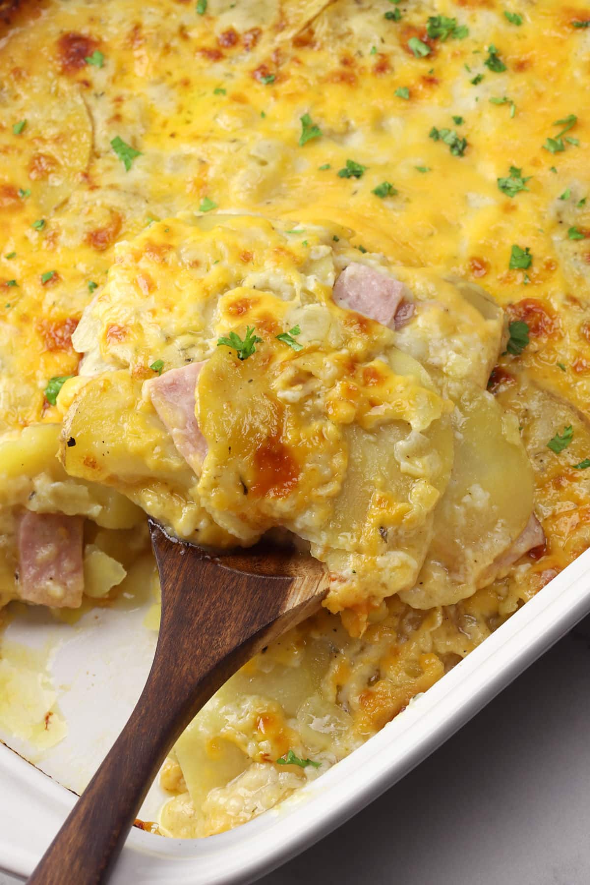 A wooden spatula scooping sliced cheesy potatoes with ham from a casserole dish.