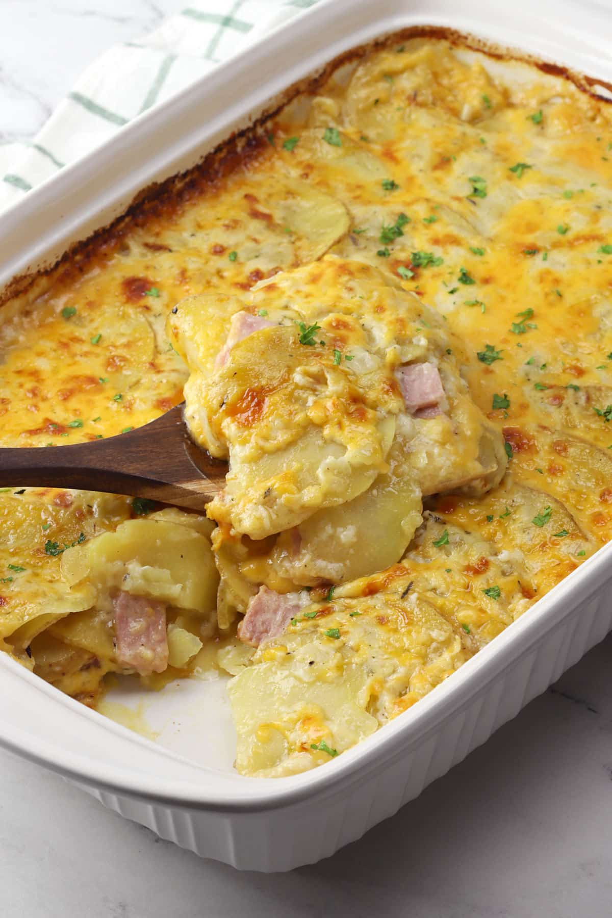 A white casserole dish filled with layers of thin sliced potatoes, ham, and cheese, with a wooden spatula scooping a serving out.