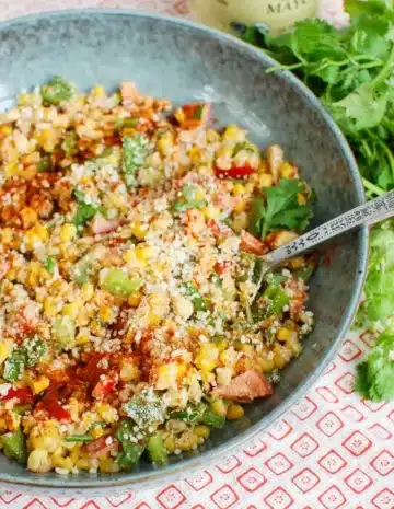 A blue bowl of Mexican corn salad with a metal serving spoon.