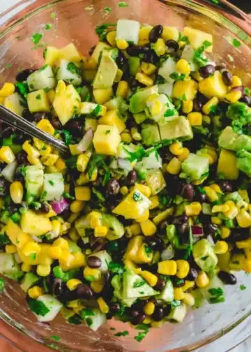 A glass bowl of mango and black bean salad with a metal serving spoon.