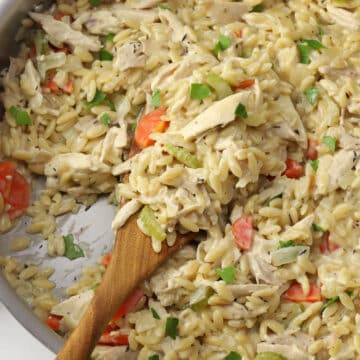 Close up of creamy chicken orzo in a skillet scooped by a wooden serving spoon.