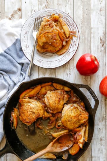 A cast iron pan and serving plate of chicken thighs and apple slices.