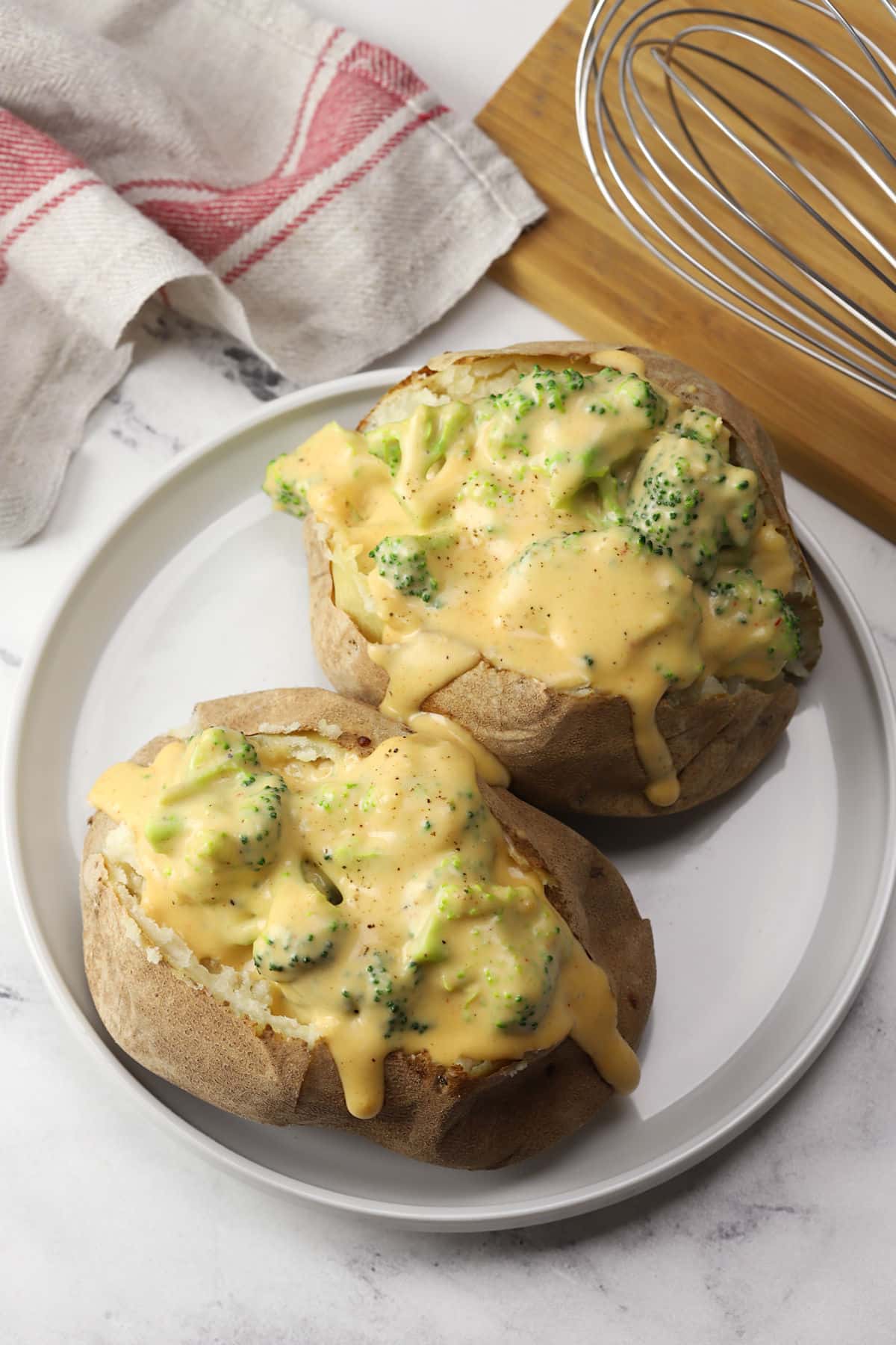 Two baked potatoes topped with broccoli cheese sauce on a white plate.