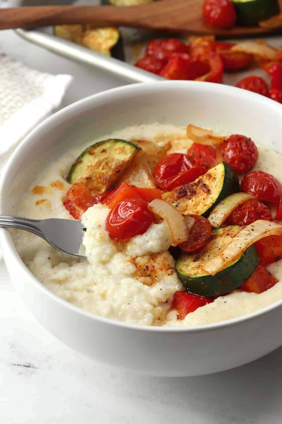 A fork in a bowl of creamy grits and roasted vegetables.