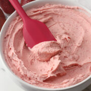 A red spatula in a bowl of pink strawberry buttercream frosting.