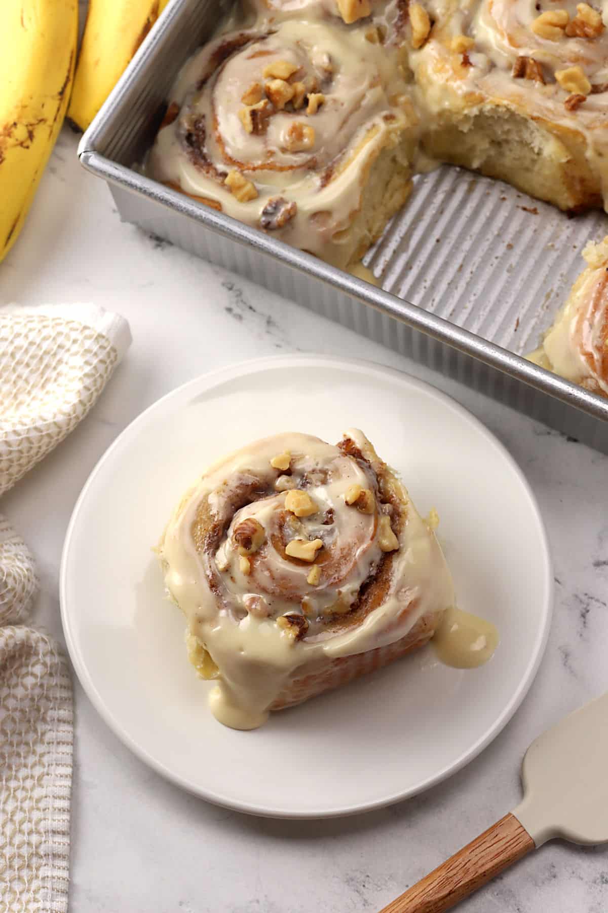 A banana bread cinnamon roll on a plate with more in a pan in the background.