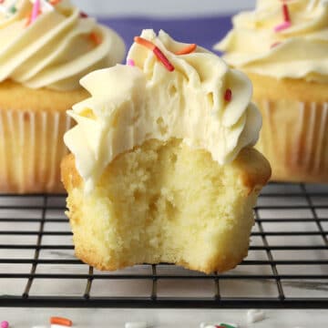 Vanilla cupcake sitting on a cooling rack with a bite missing.