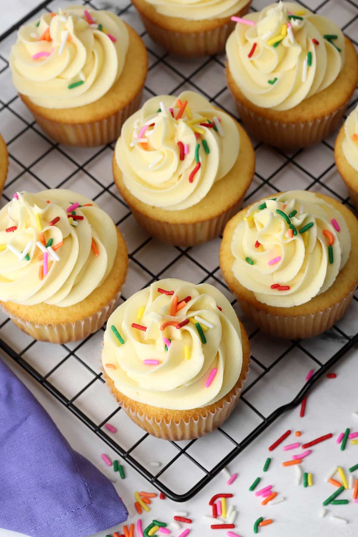 Vanilla cupcakes on a cooling rack topped with frosting and sprinkles.