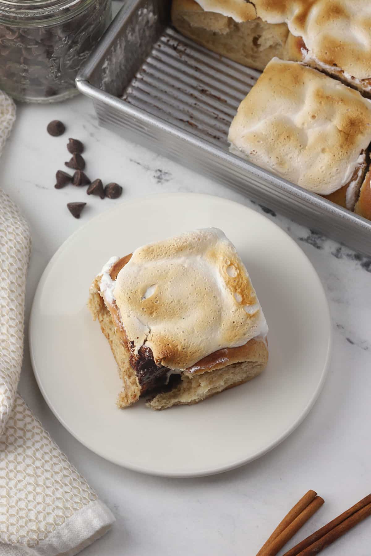 A cinnamon roll on a plate with a pan of more in the background.