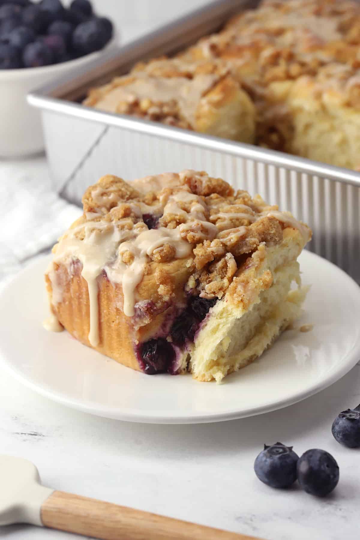 A cinnamon roll on a plate with a pan of more in the background.