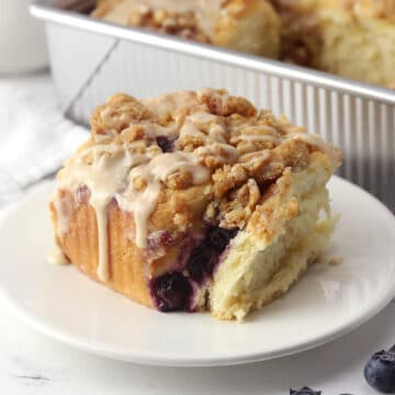 A cinnamon roll on a plate with a pan of more in the background.