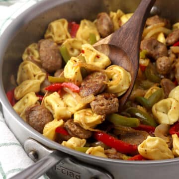 A pan of tortellini, sausage, and vegetables being scooped with a wooden spatula.