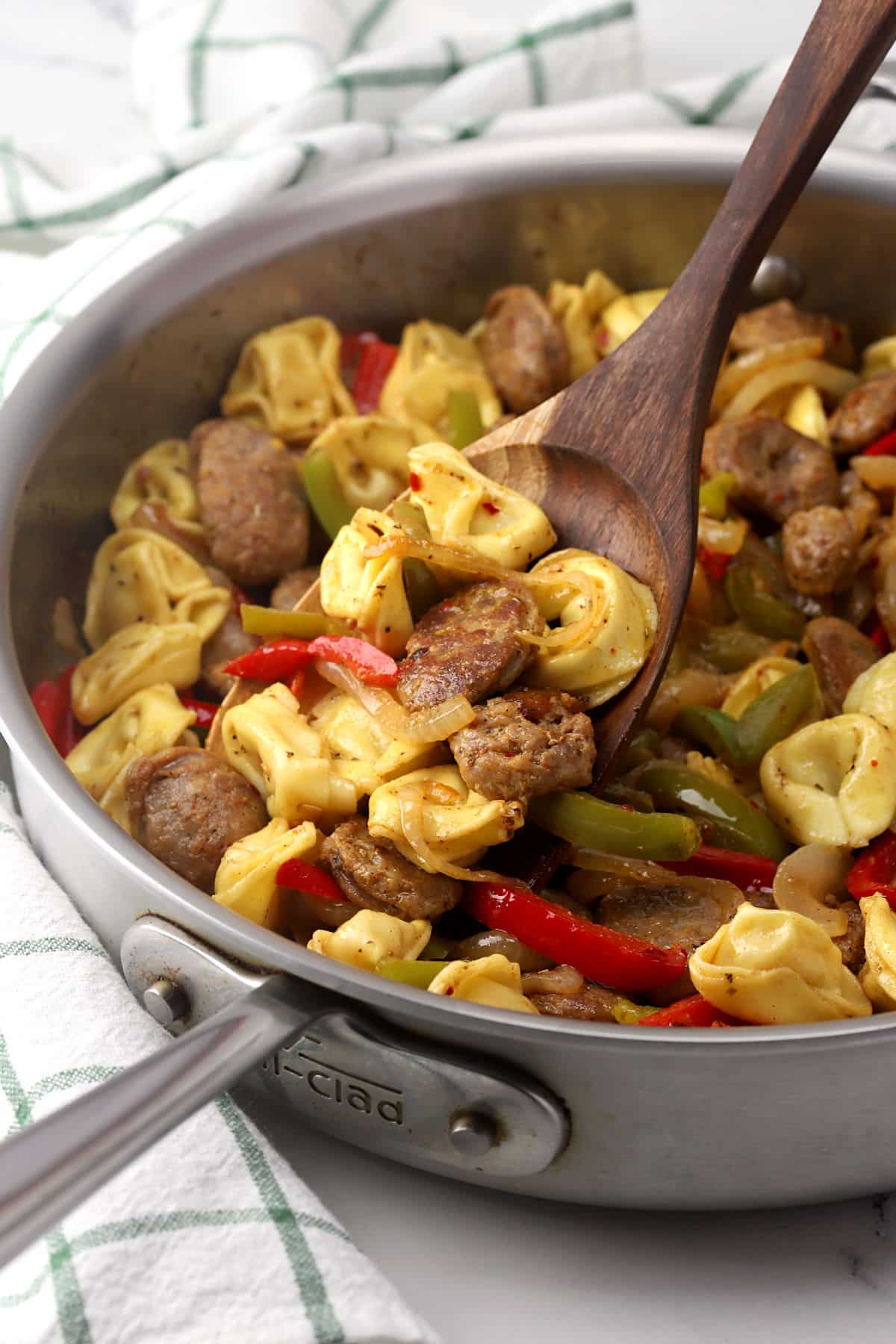 A pan of tortellini, sausage, and vegetables being scooped with a wooden spatula.