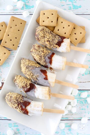 A serving platter of chocolate covered frozen bananas on sticks with graham crackers for garnish.