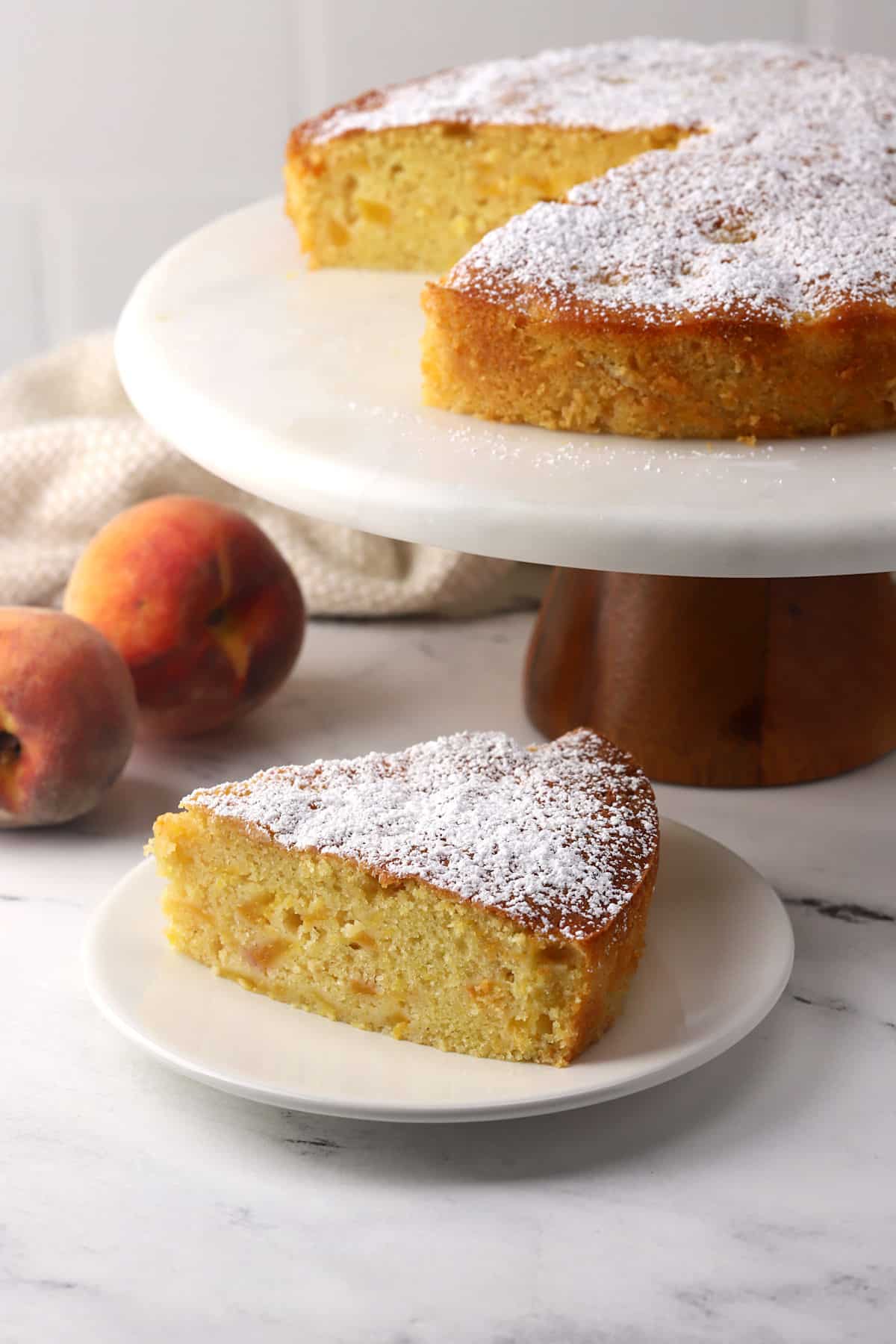 A slice of peach cake on a small plate with the whole cake on a cake stand behind.