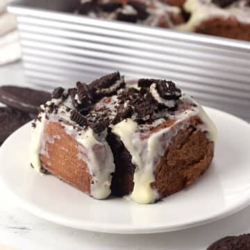 A cookies and cream cinnamon roll on a plate with a pan of others in the back.