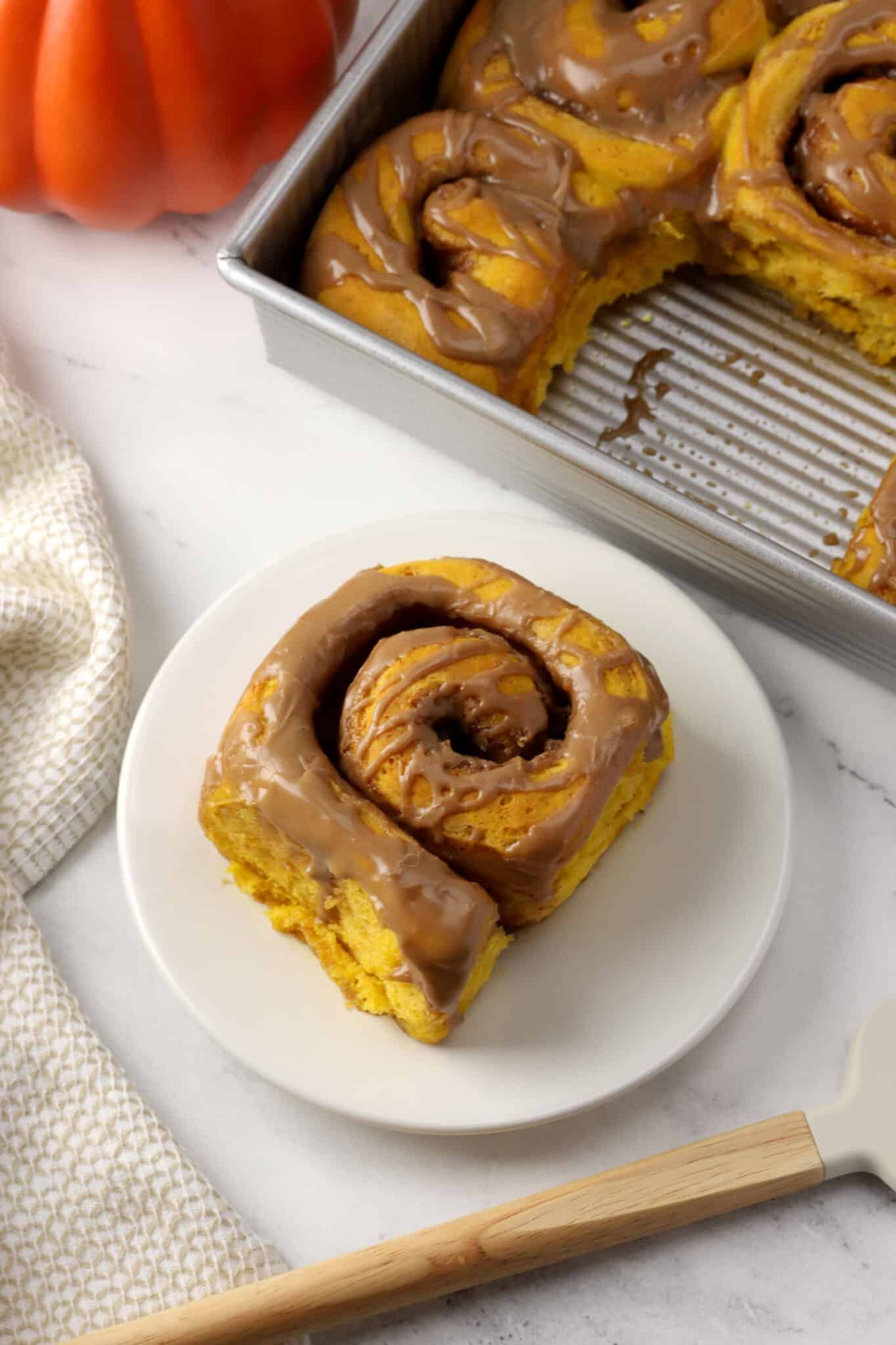 A pumpkin chai cinnamon roll on a small plate with a pan of cinnamon rolls in the back ground.