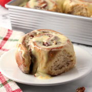 A cinnamon roll on a small plate with a pan of more in the background.