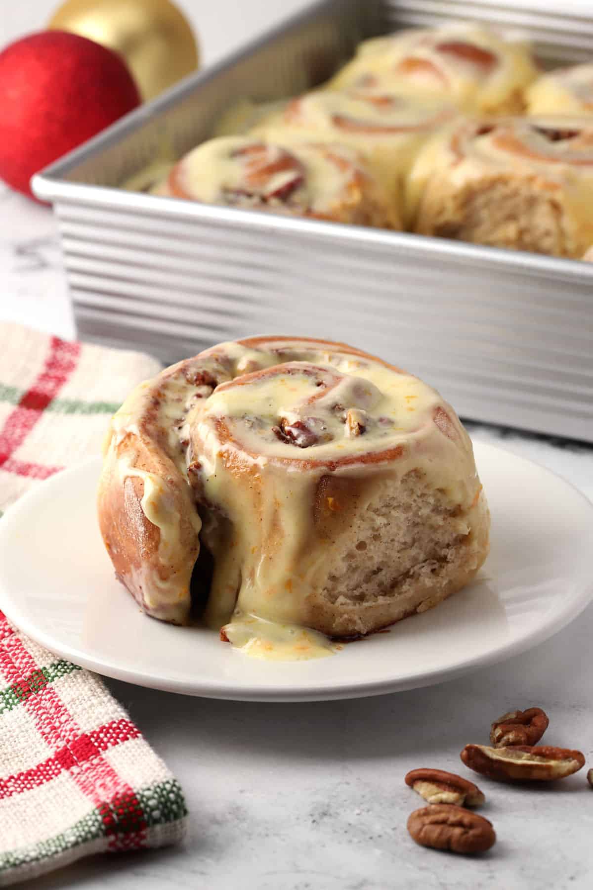 A cinnamon roll on a small plate with a pan of more in the background.