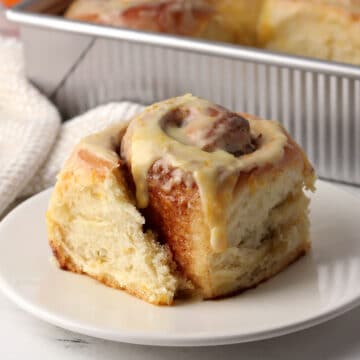 A cinnamon roll on a small plate with a pan of more in the back ground.