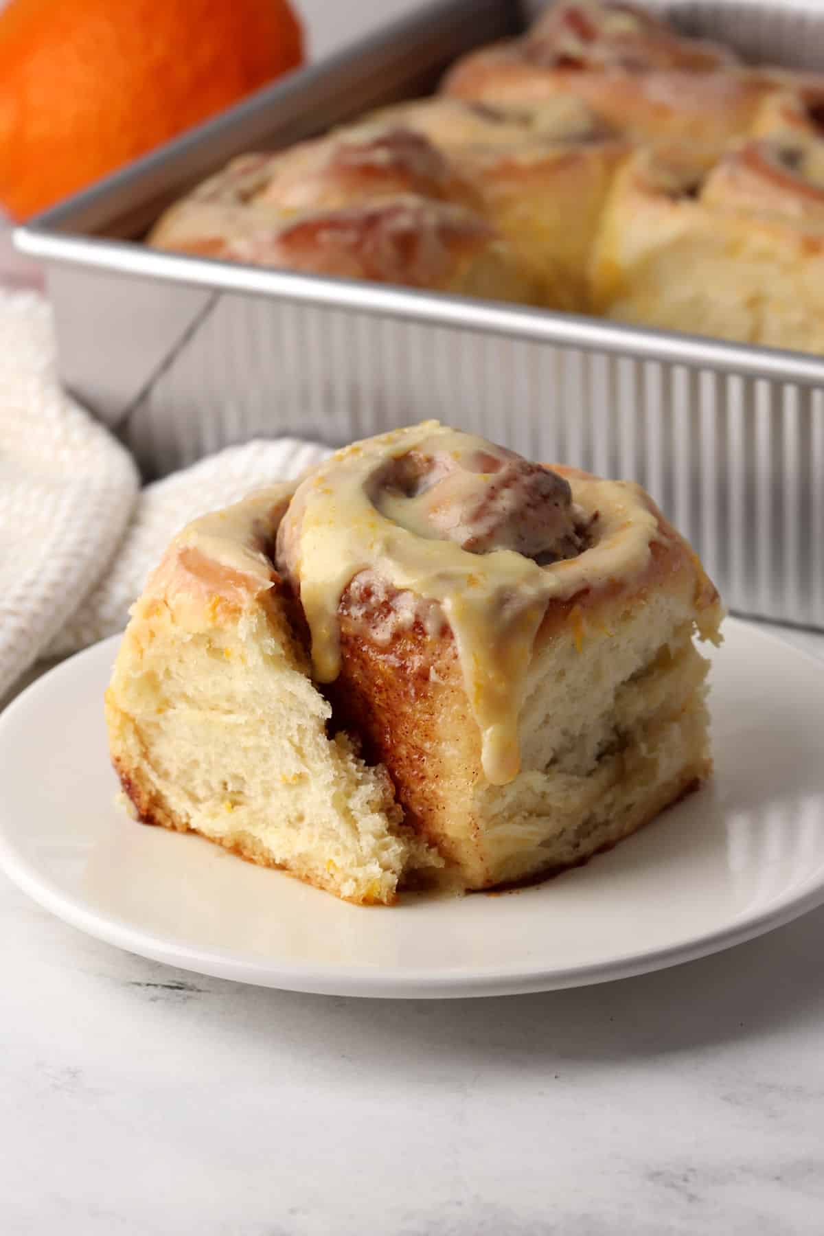 A cinnamon roll on a small plate with a pan of more in the back ground.