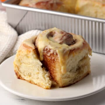 A cinnamon roll on a small plate with a pan of more in the back ground.