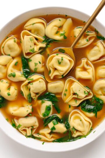 A bowl of broth and tortellini with a spoon.